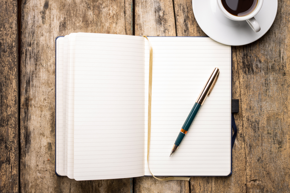 ett block och en penna på ett träbord med en kaffekopp i det högra hörnet. Ett anteckningsblock för att skriva upp sitt mål i